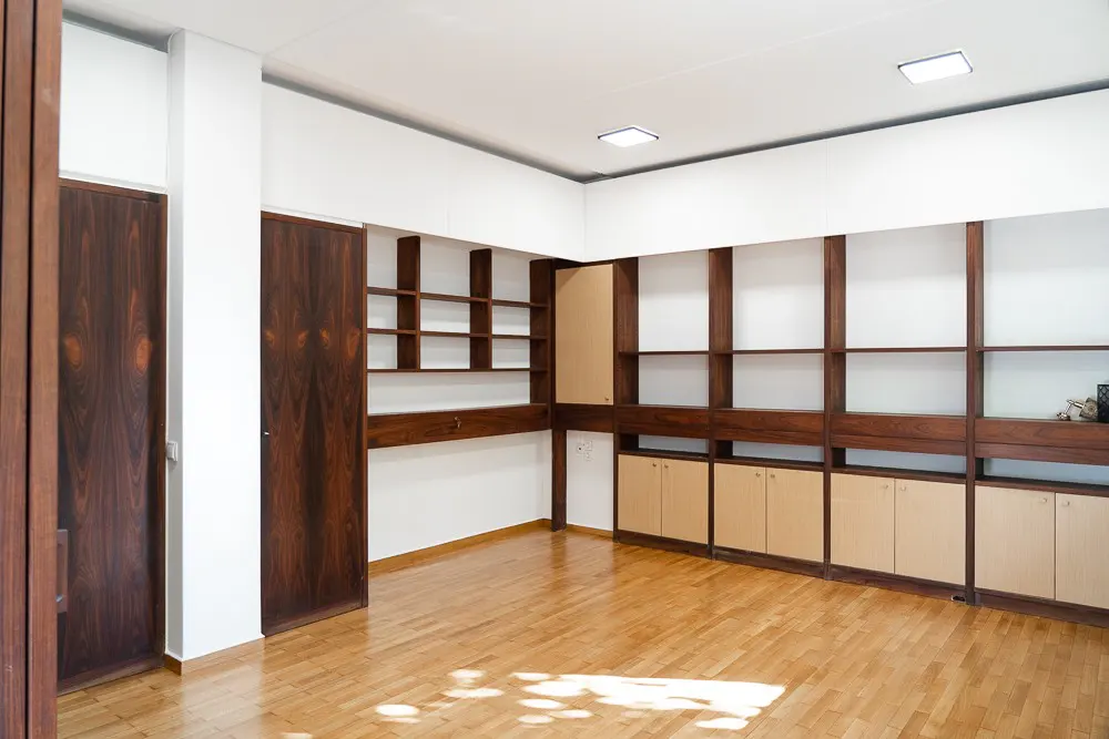 Bright room with wood floors, white walls, and built-in shelving. Dark wood shelves and cabinets line the back wall. Two dark wood doors are on the left.