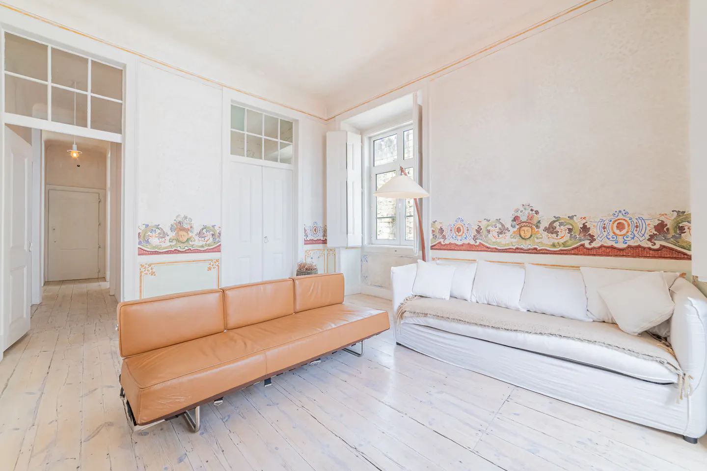 Bright living room with white walls, painted trim, and white wood floors. A tan leather sofa and a white sofa with pillows are present.