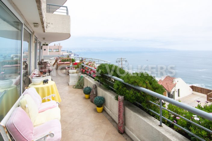 A wide balcony with pink and yellow furniture overlooks the ocean on a cloudy day. [AI-generated text]