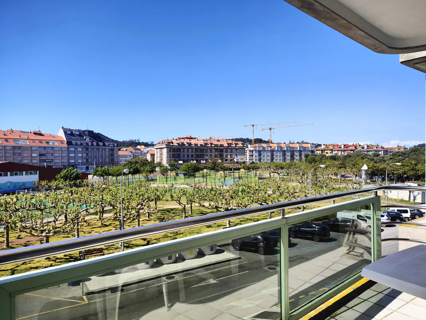 Balcony view of a city park with buildings, trees, and cars under a clear blue sky.