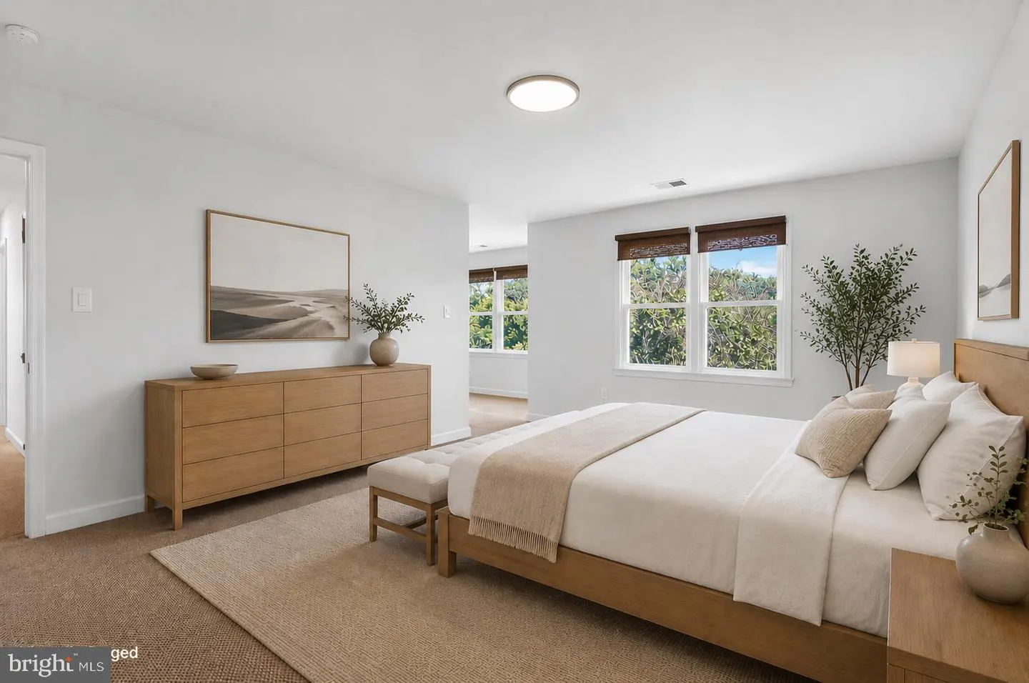 Bright bedroom with a wooden bed, white bedding, a long dresser, and windows overlooking green trees.