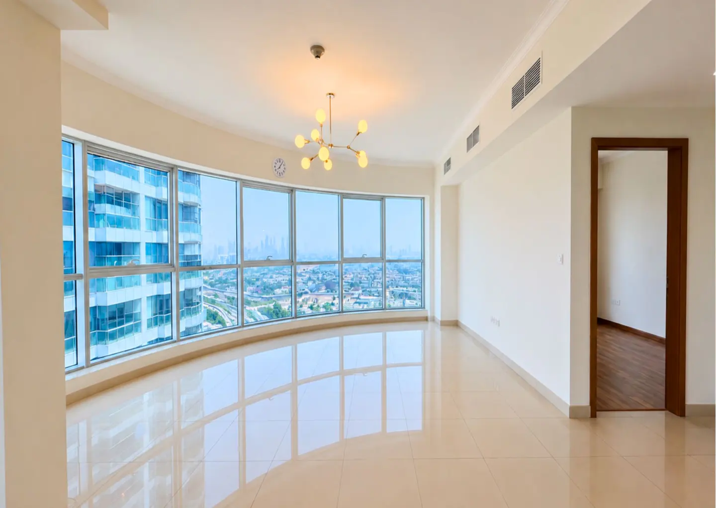 Bright, empty living room with beige tile floors, a modern chandelier, and floor-to-ceiling windows with a city view.