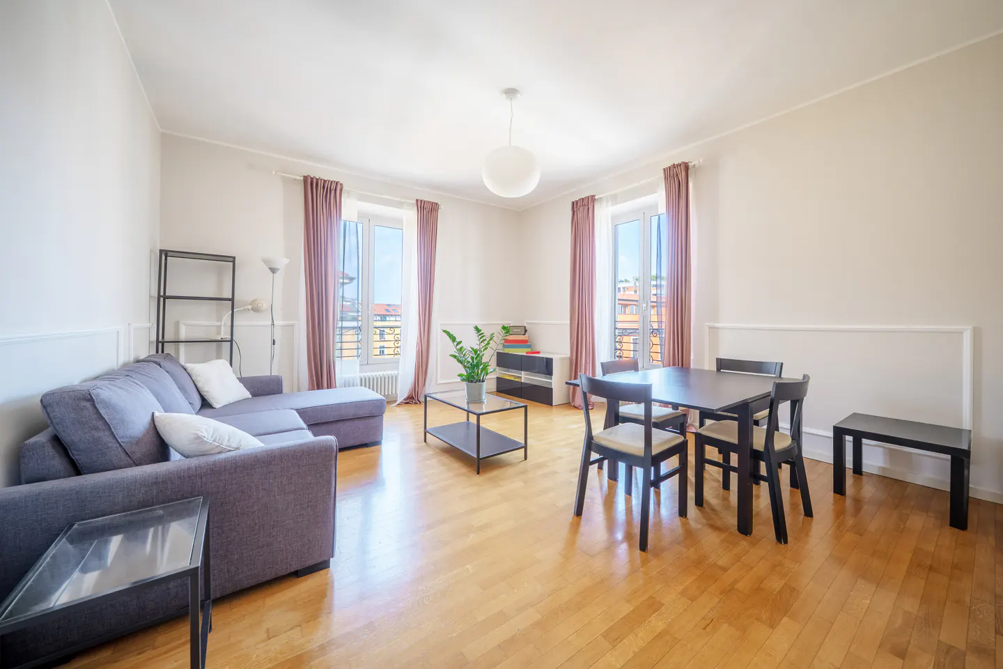 Bright living room with hardwood floors, a gray sofa, a black dining table with chairs, and windows with pink curtains.