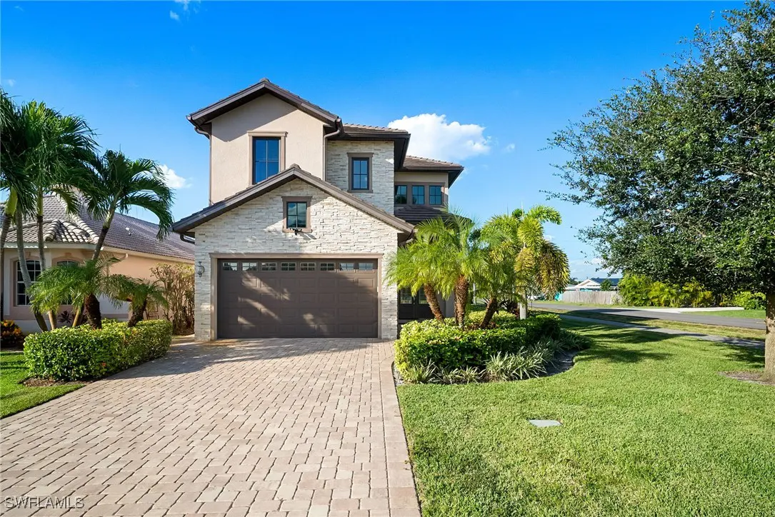 Two-story house with a brown garage door, stone accents, and a brick driveway under a blue sky. Palm trees and green grass surround the property.