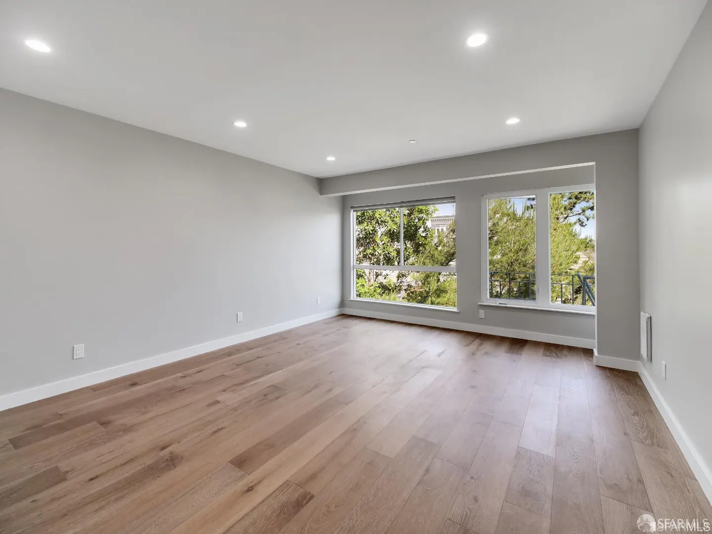 Bright, empty room with light gray walls, wood floors, and large windows showing green trees outside.