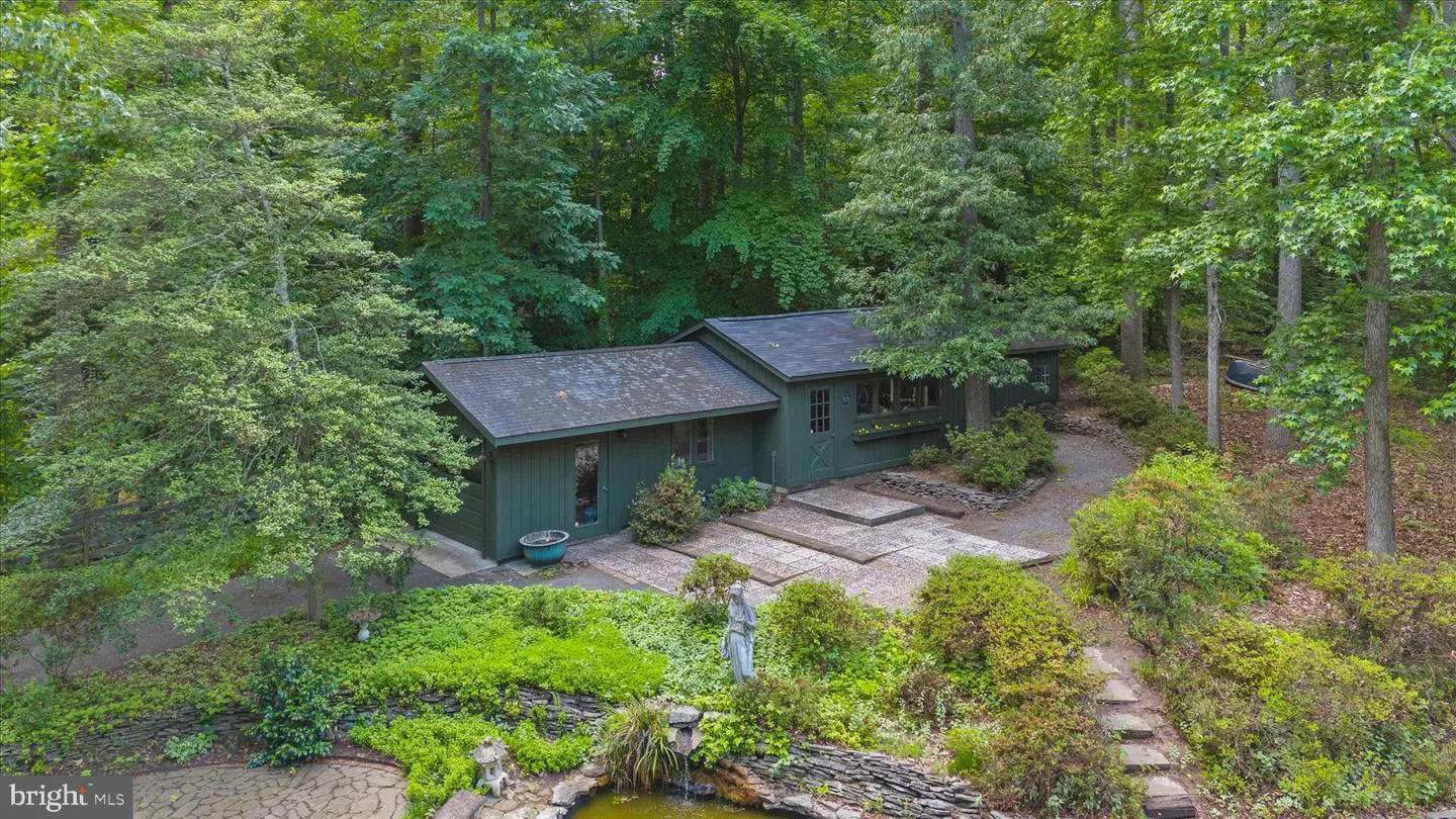 A dark green cottage nestled in a lush, wooded landscape with a stone patio, garden statue, and small pond.