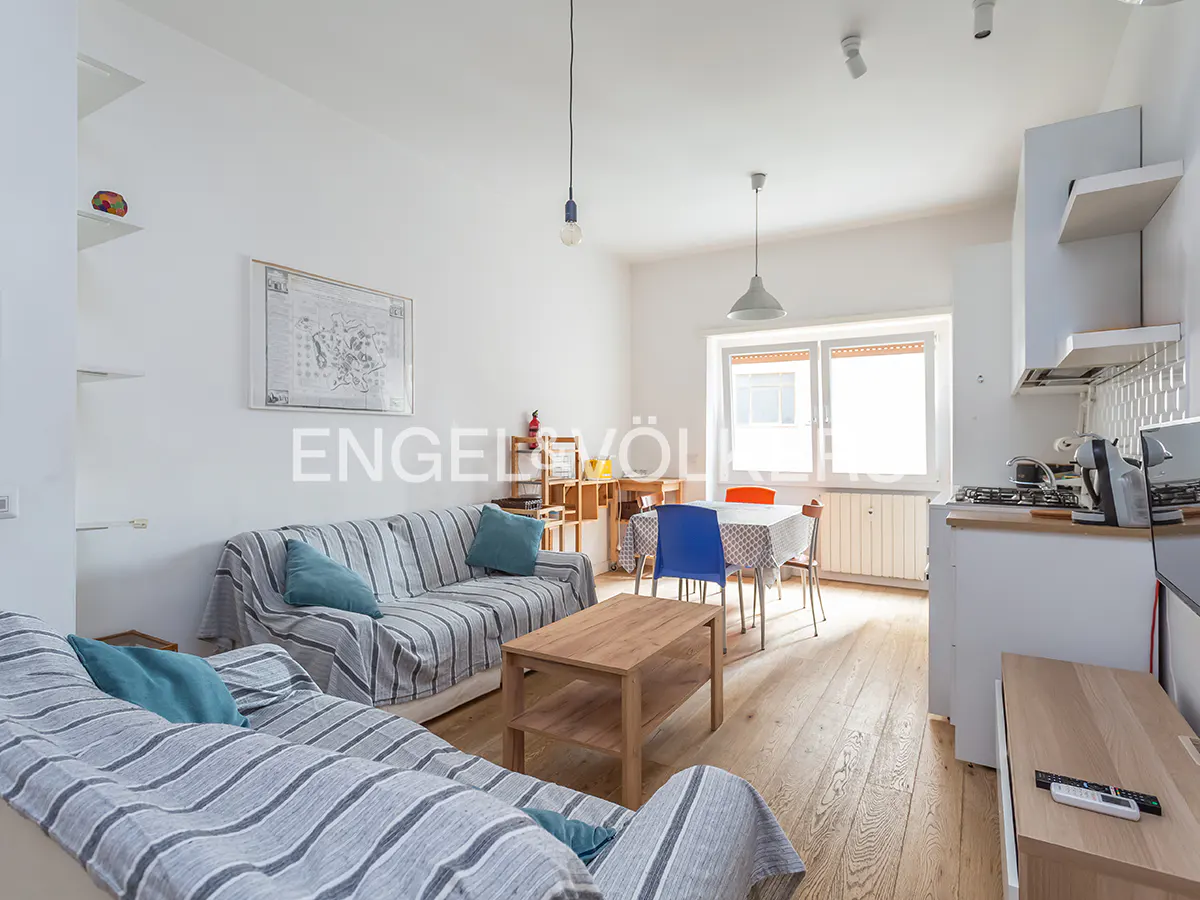 Bright living room with wood floors, two sofas with blue pillows, a dining table, and a small kitchen area.