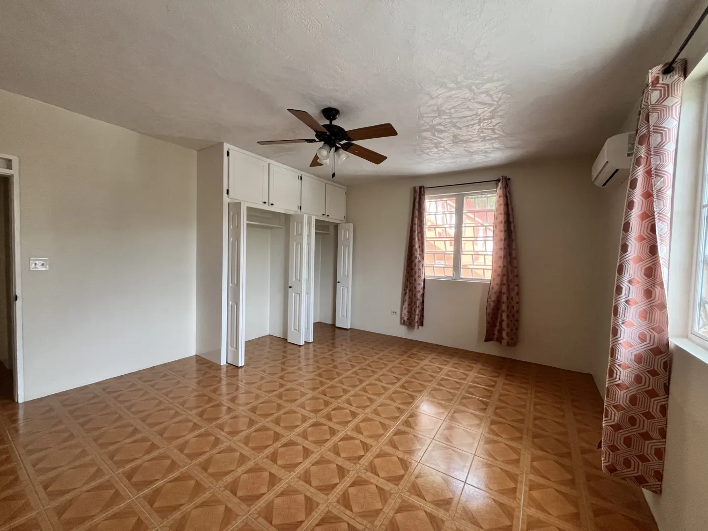 Empty bedroom with beige walls, patterned tile floors, white built-in closets, a ceiling fan, and patterned curtains.
