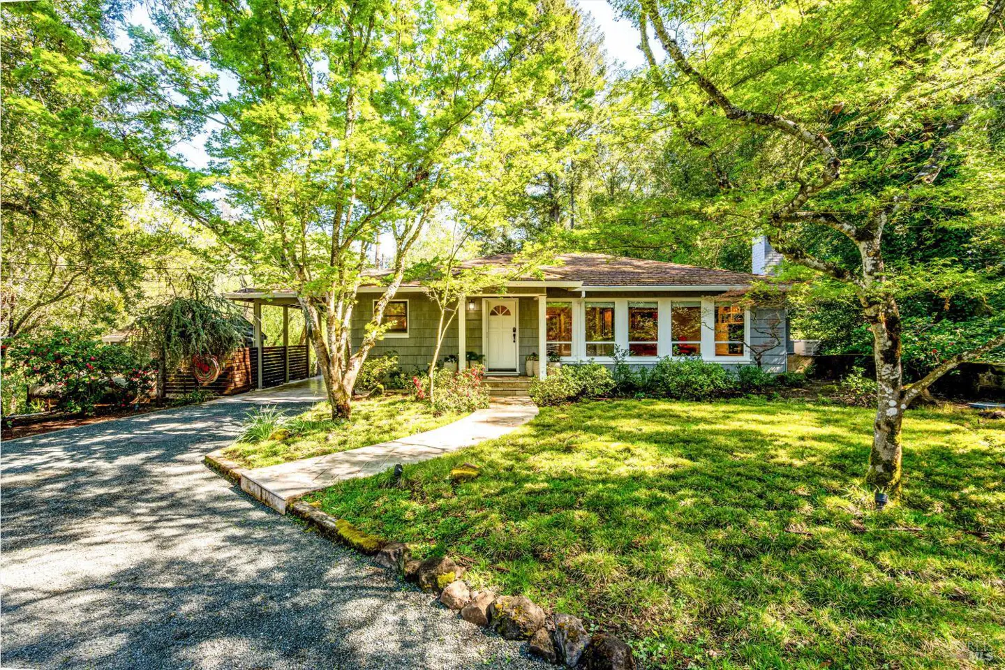A single-story gray house with a white door and trim, surrounded by green trees and lawn.