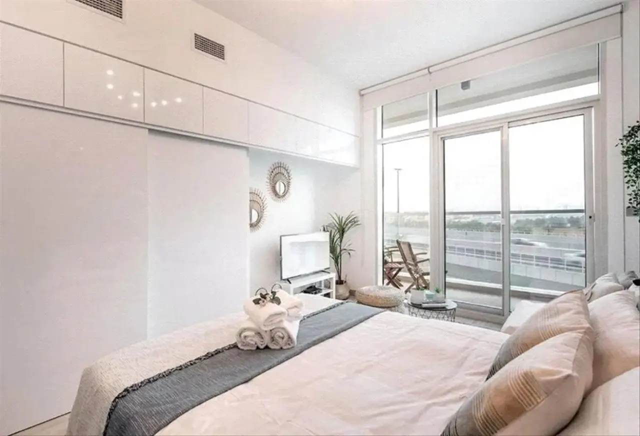 Bright bedroom with white walls, cabinets, and bedding. A sliding glass door leads to a balcony with chairs. Towels are neatly stacked on the bed.