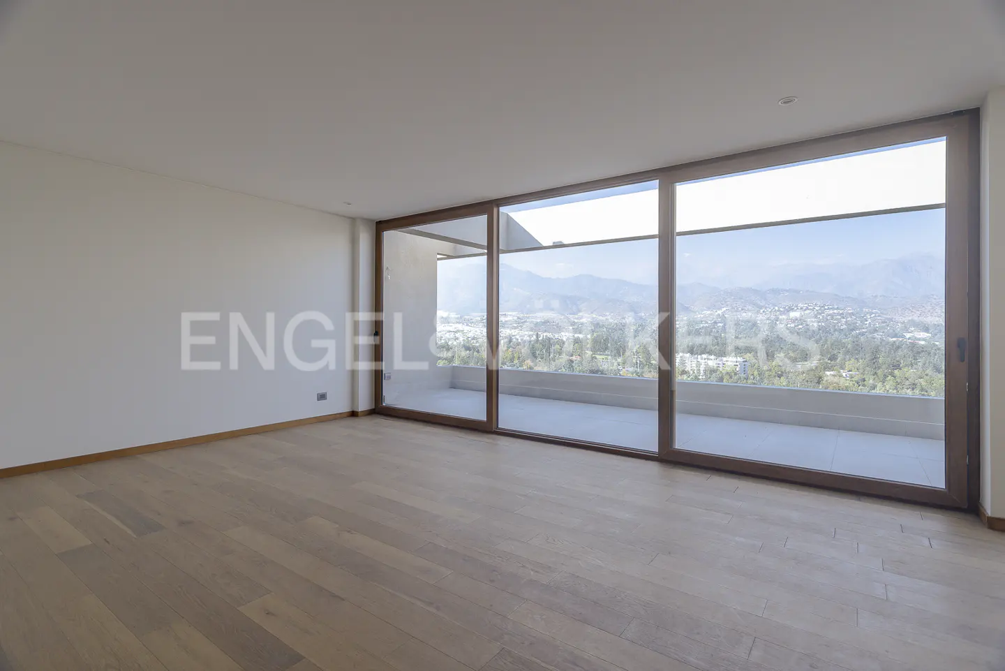 Empty room with light wood floors and white walls. Large windows offer a view of a city and mountains.