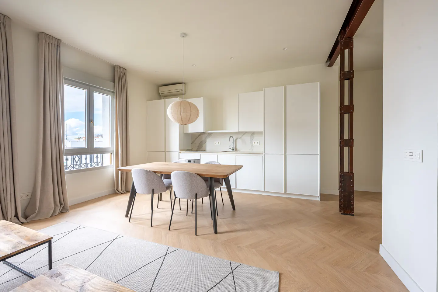 Bright apartment with herringbone floors, white kitchen cabinets, and a wooden dining table with four gray chairs. A large window with curtains is on the left.