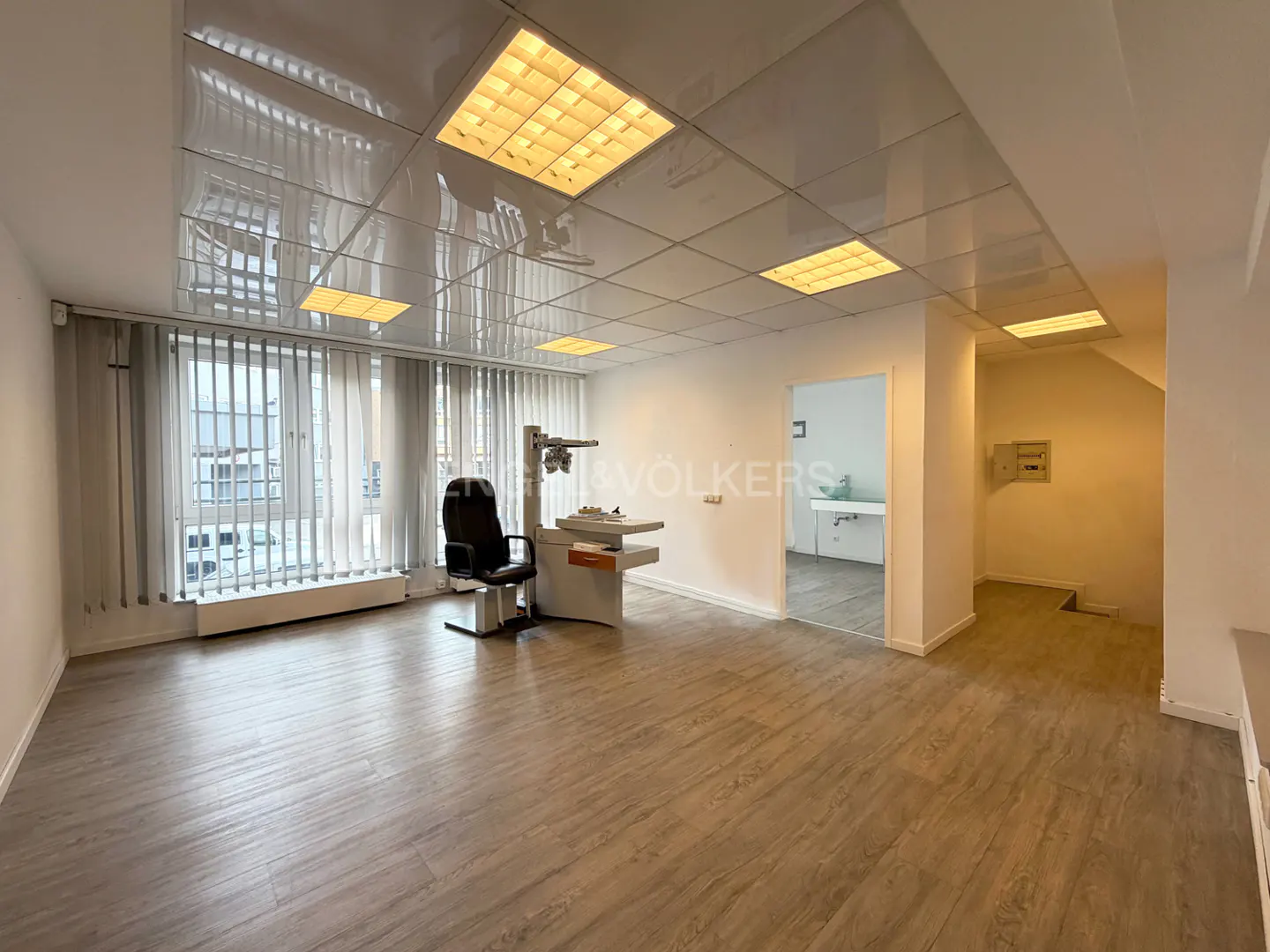 Bright, empty office space with wood floors, white walls, and a drop ceiling with fluorescent lights. An eye exam machine and chair sit near a window.