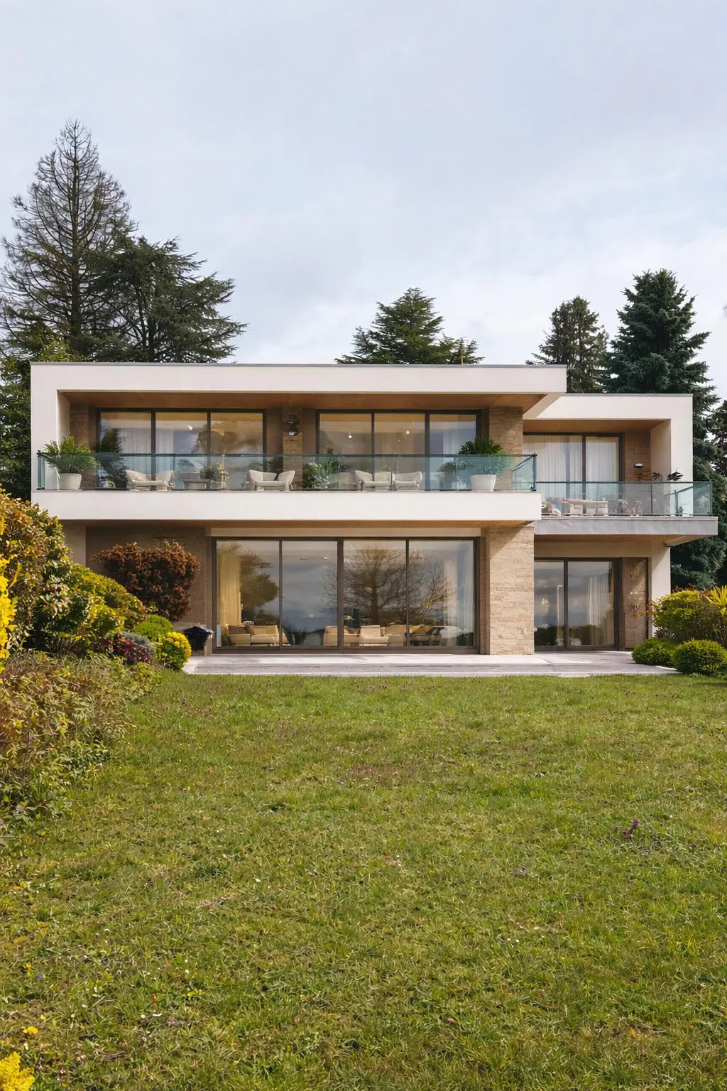 Two-story modern house with large windows and balconies overlooking a green lawn and trees.