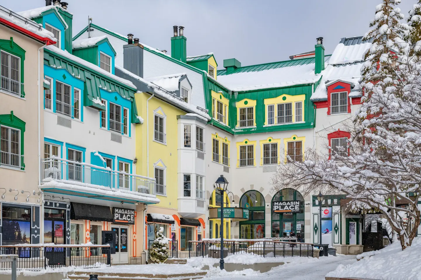 Colorful buildings with snow-covered roofs line a street in a winter resort town. Shops are visible on the ground floor.
