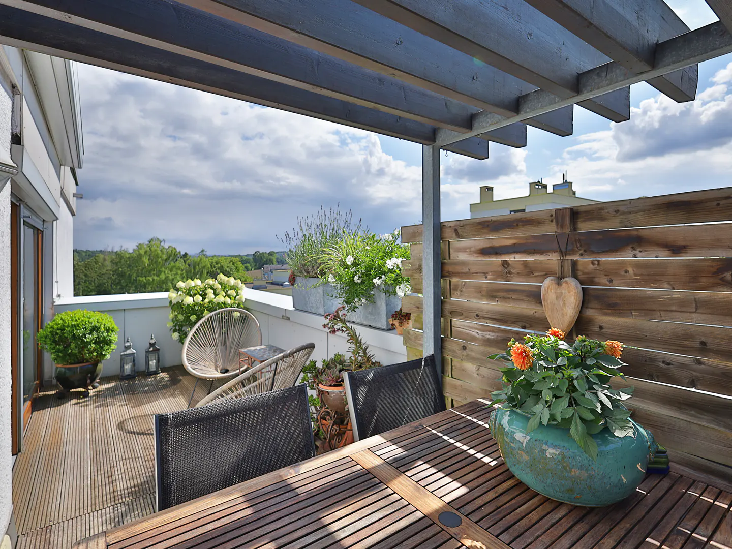 A rooftop patio with a wooden table, chairs, and potted plants, under a pergola with a view of trees and cloudy sky.