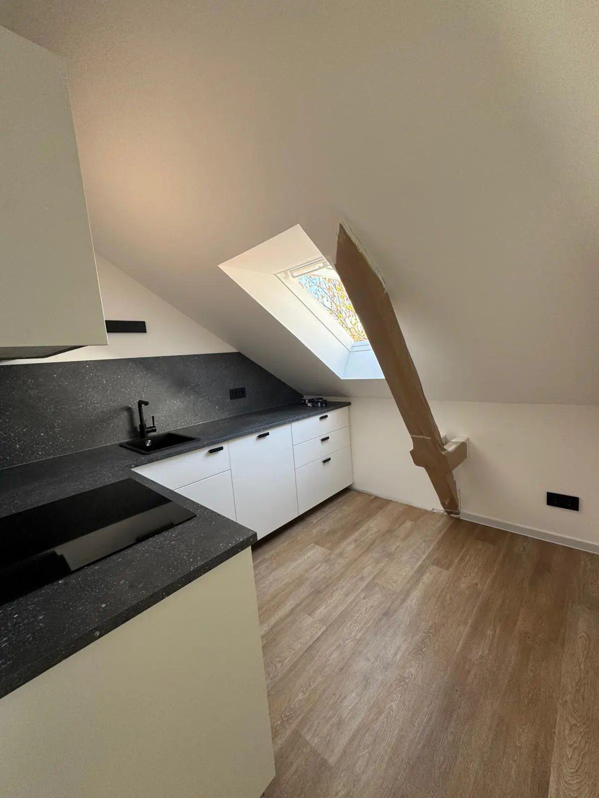 Attic apartment kitchen with white cabinets, black countertops, and wood floors. A skylight and exposed beam add character.
