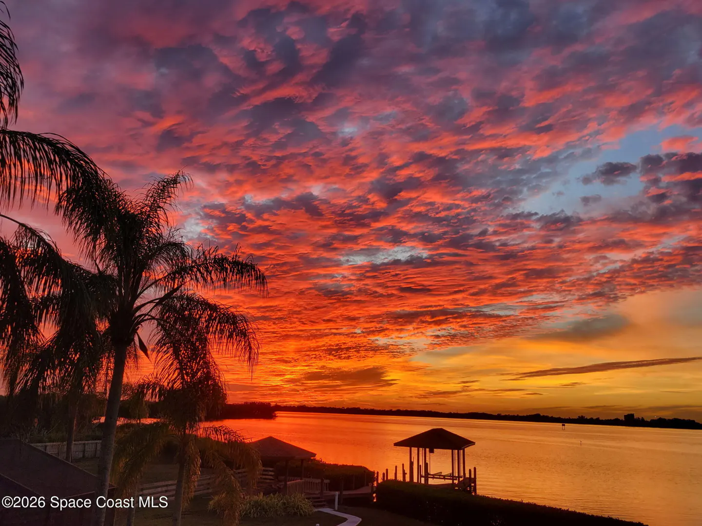A vibrant sunset over water, with palm trees silhouetted on the left. The sky is filled with red and orange clouds. Two gazebos are visible on the shoreline.