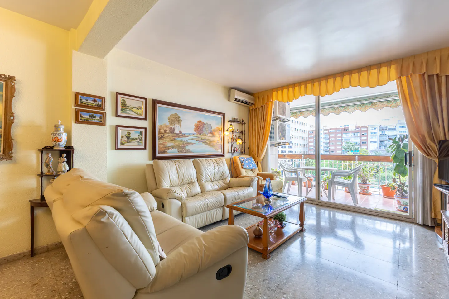 Living room with cream leather sofas, wood coffee table, and balcony with city view. Paintings adorn the yellow walls.