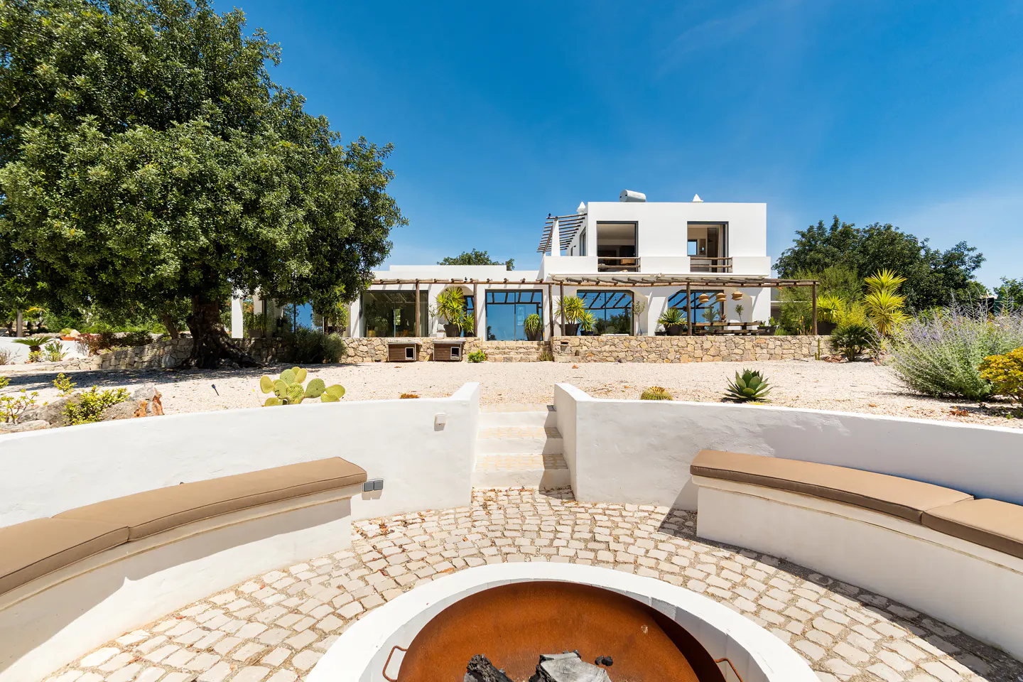 A white villa with a stone patio, large tree, and a circular fire pit area with curved benches in the foreground.
