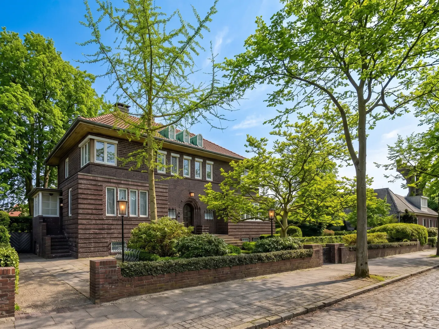 Two-story brick house with white-framed windows, surrounded by green trees and bushes, under a blue sky.