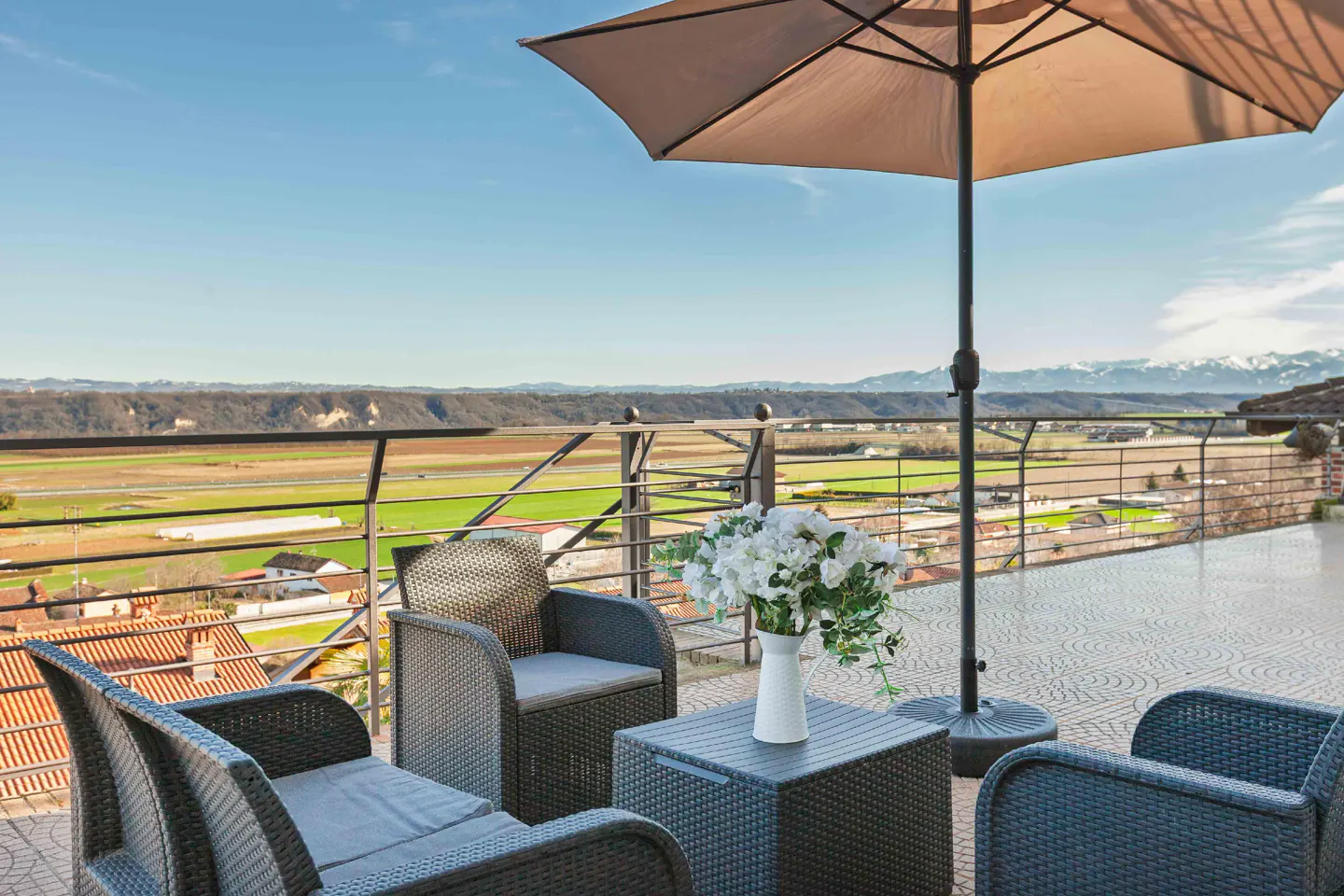 Outdoor patio with wicker furniture, umbrella, and white flowers overlooking a green valley and distant mountains.