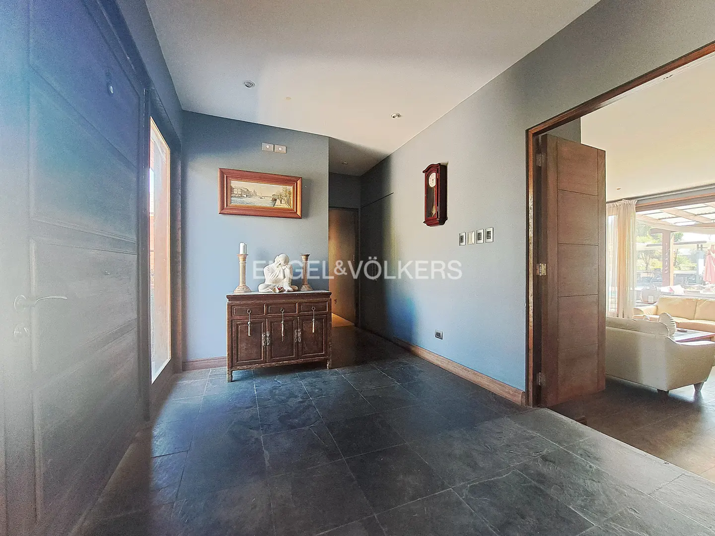 Entryway with slate floors, gray walls, and a dark wood cabinet topped with candles and a sculpture. A doorway leads to a bright living room.