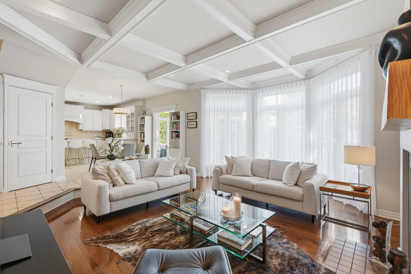 Bright living room with two white sofas, a glass coffee table on a cowhide rug, and an open-concept kitchen in the back.