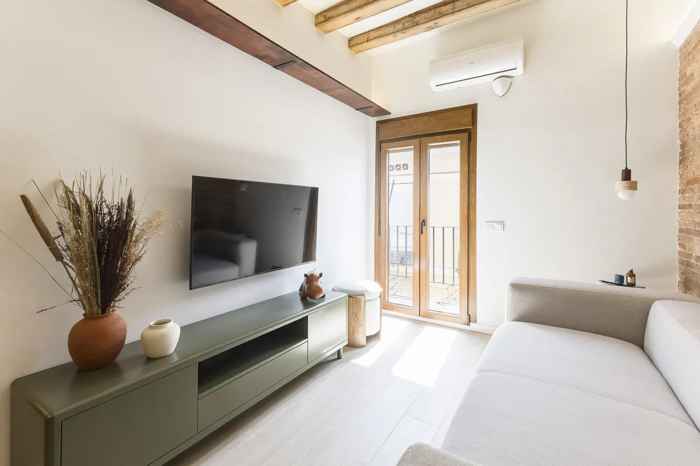 Living room with white walls, wooden beams, and a TV above a green cabinet. A white sofa sits near a balcony door.