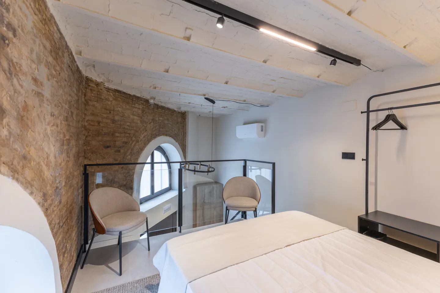 Bedroom with white linens, exposed brick wall, arched window, two chairs, and black clothing rack.