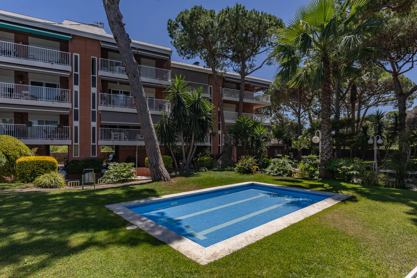 Rectangular swimming pool in a lush green garden with a multi-story apartment building and tall pine trees in the background.