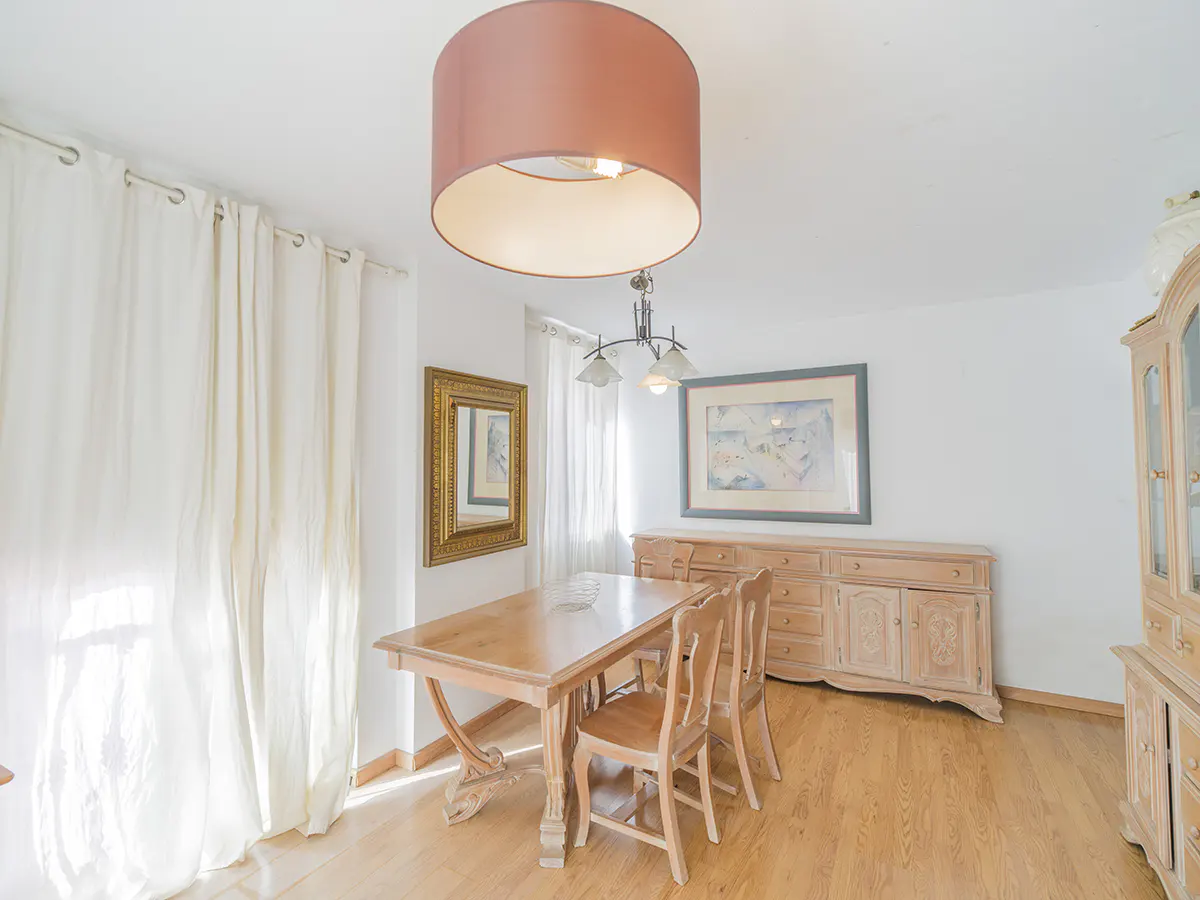 Bright dining room with wood table, chairs, and floor. A large, round, pink lampshade hangs above. White curtains and walls create a clean, airy feel.