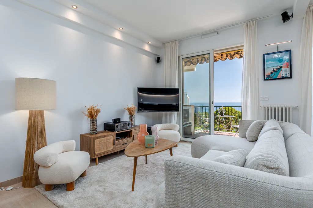 Bright living room with a white sofa, cream armchair, wooden media console, and a balcony view of the ocean.