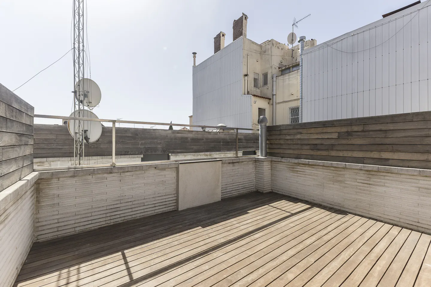 A rooftop terrace with wooden decking, a brick and wood perimeter wall, and buildings in the background.