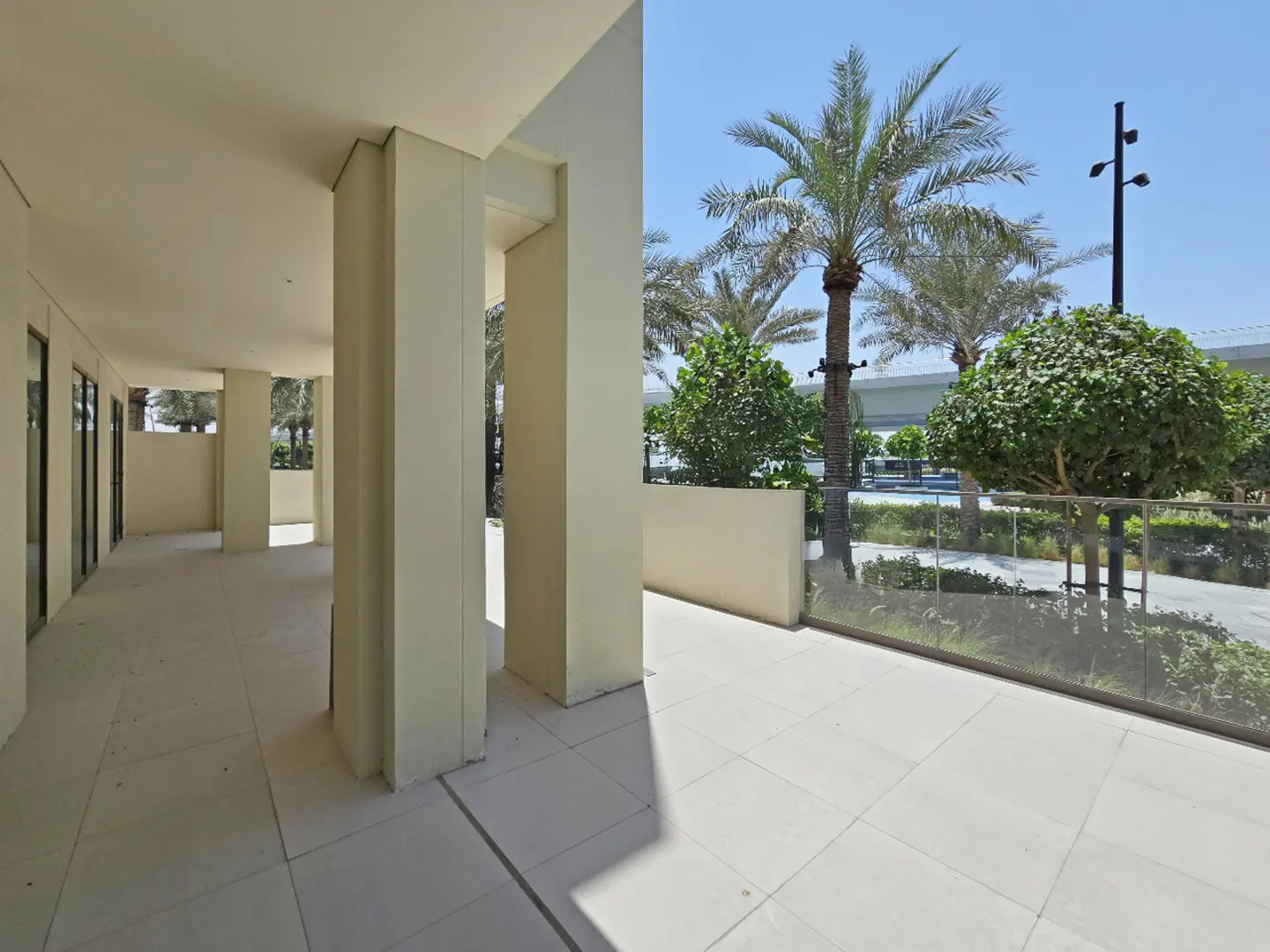 Outdoor patio with cream-colored columns, tiled floor, and glass railing. Palm trees and greenery are visible beyond the railing.