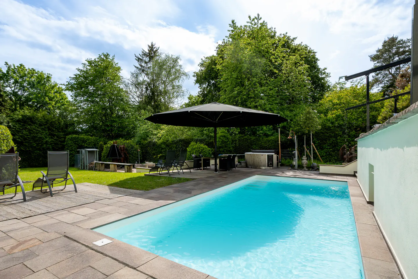 A rectangular swimming pool with turquoise water sits on a stone patio, surrounded by lounge chairs and a large umbrella.