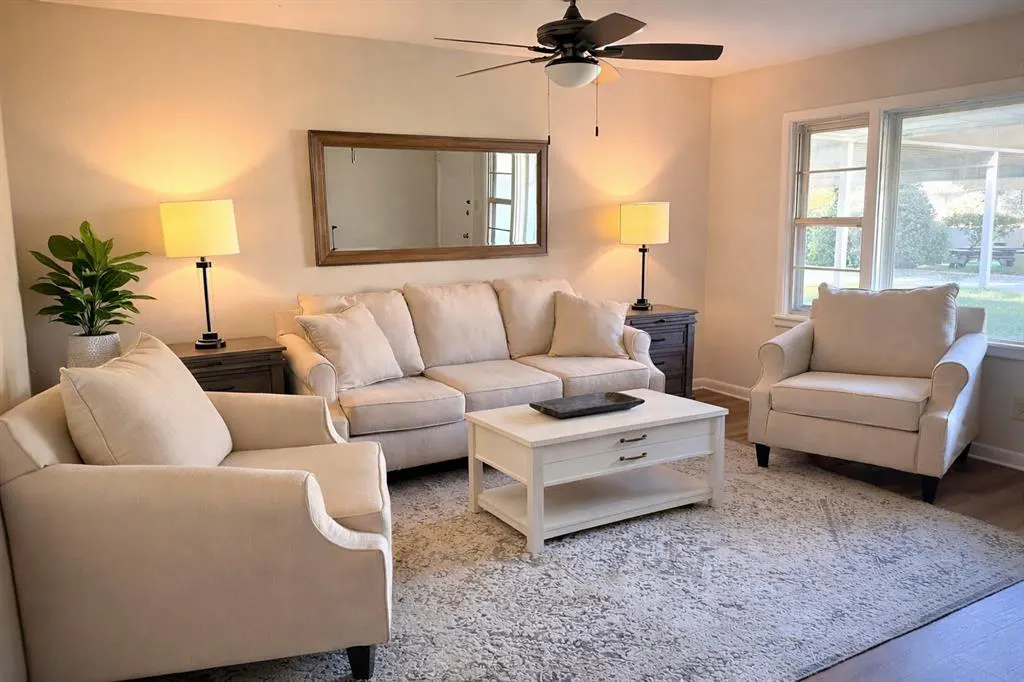 Living room with beige sofa, two armchairs, white coffee table, and patterned rug. A ceiling fan and lamps provide light.