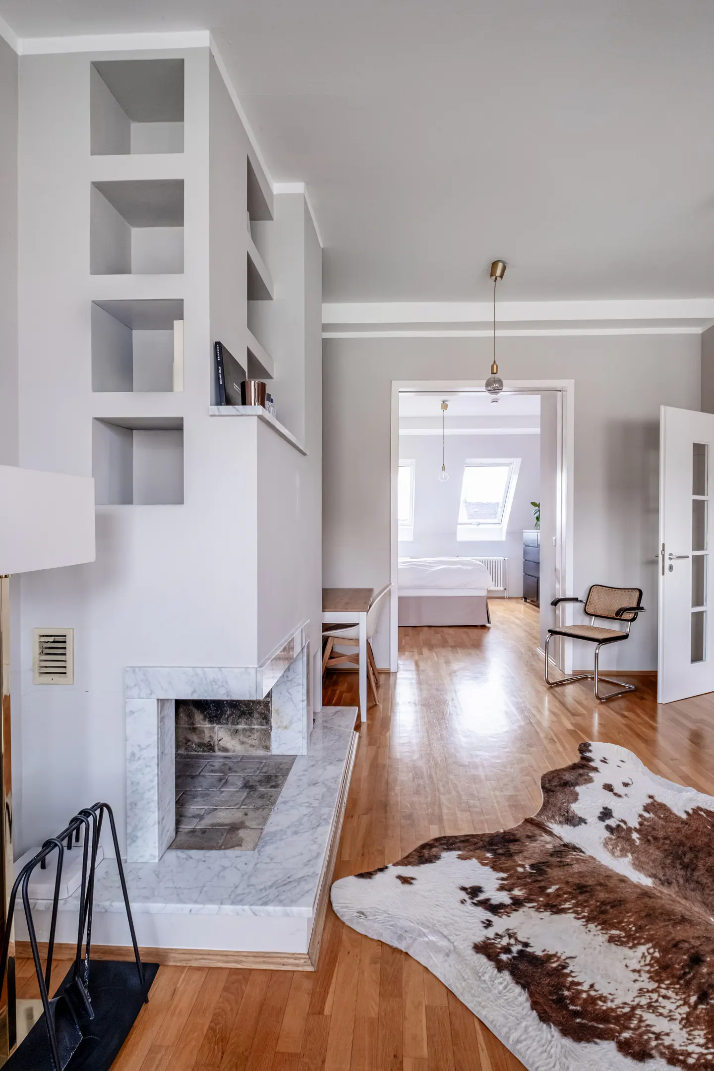A bright, modern living room with a marble fireplace, built-in shelves, and a cowhide rug on hardwood floors. A bedroom is visible through an open doorway.