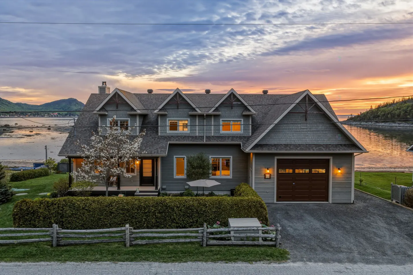Grey shingled house with a brown garage at sunset, featuring a blooming tree, hedge, and waterfront view.