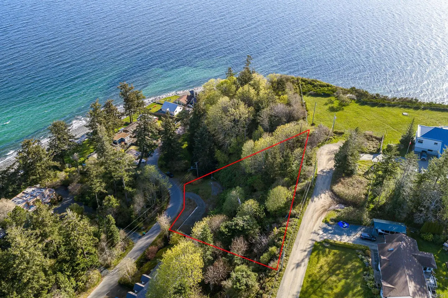 Aerial view of a wooded coastal lot outlined in red, situated between a road and the blue ocean shoreline.