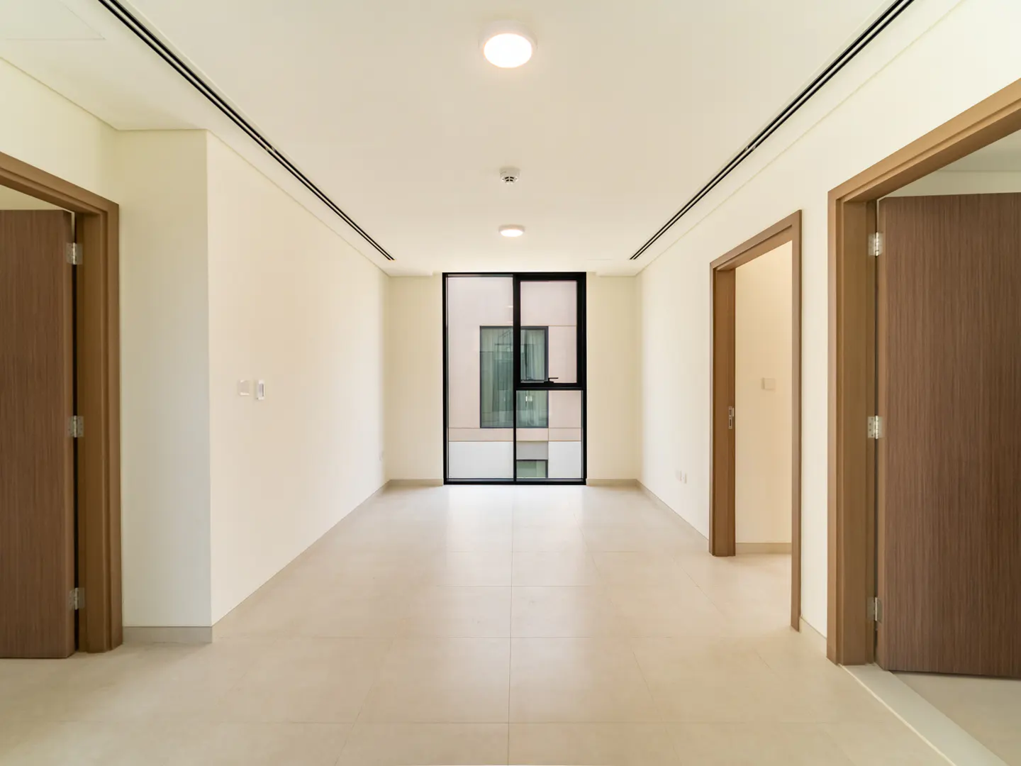 Empty room with beige tile floor, white walls, and three wood doors. A black-framed window is at the end of the room.