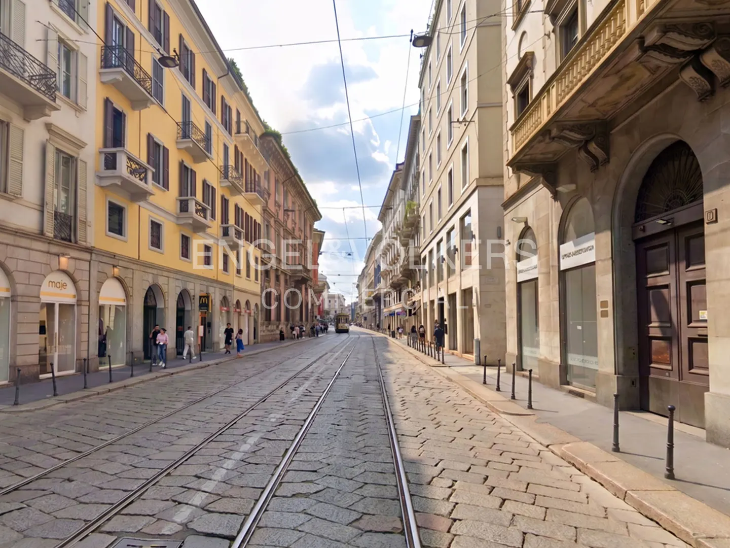 Street view of Milan, Italy, with tram tracks running down the center of a cobblestone road lined with shops and buildings.