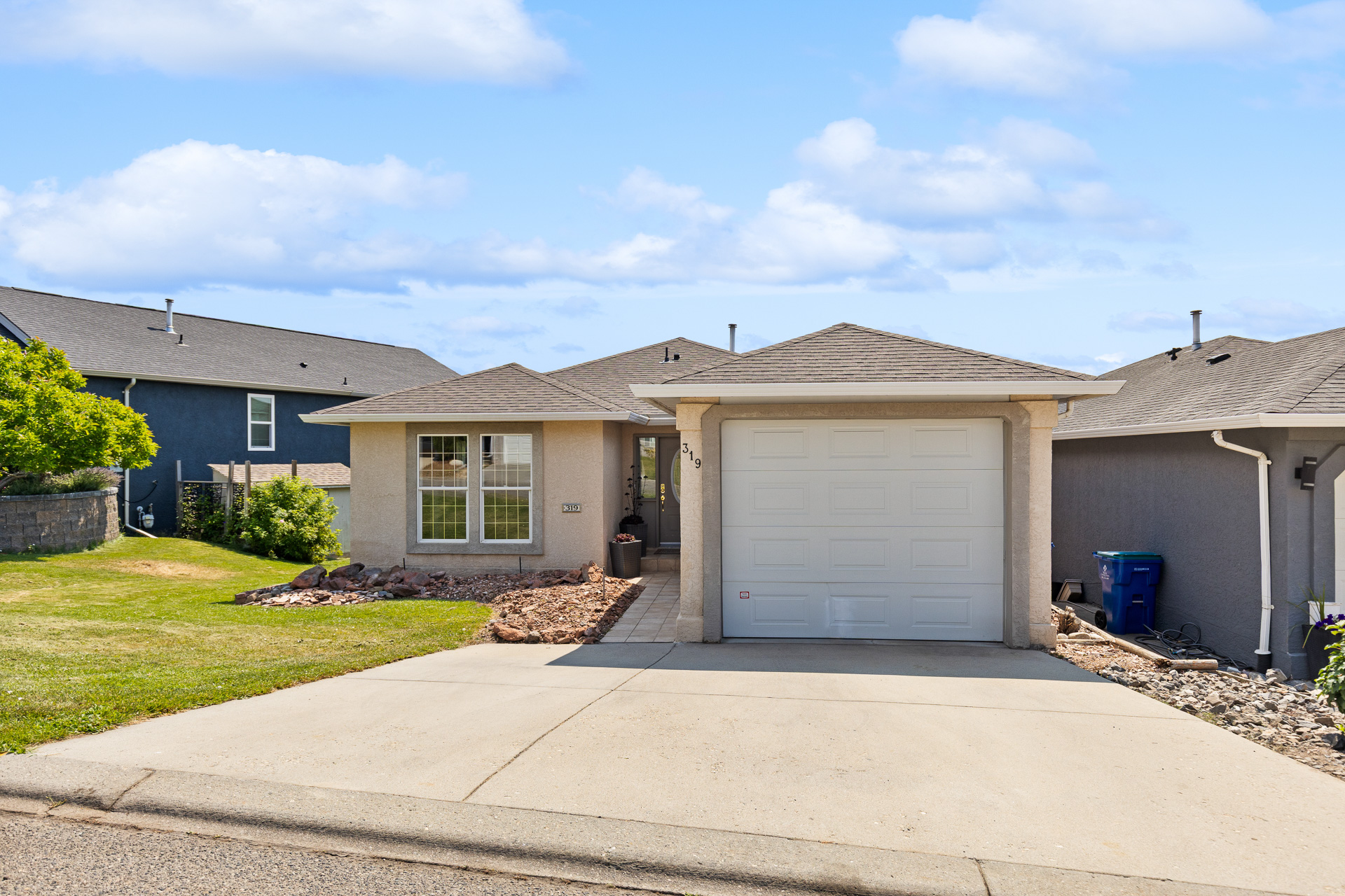 Single-story beige house with a white garage door, concrete driveway, and landscaped front yard under a blue sky.