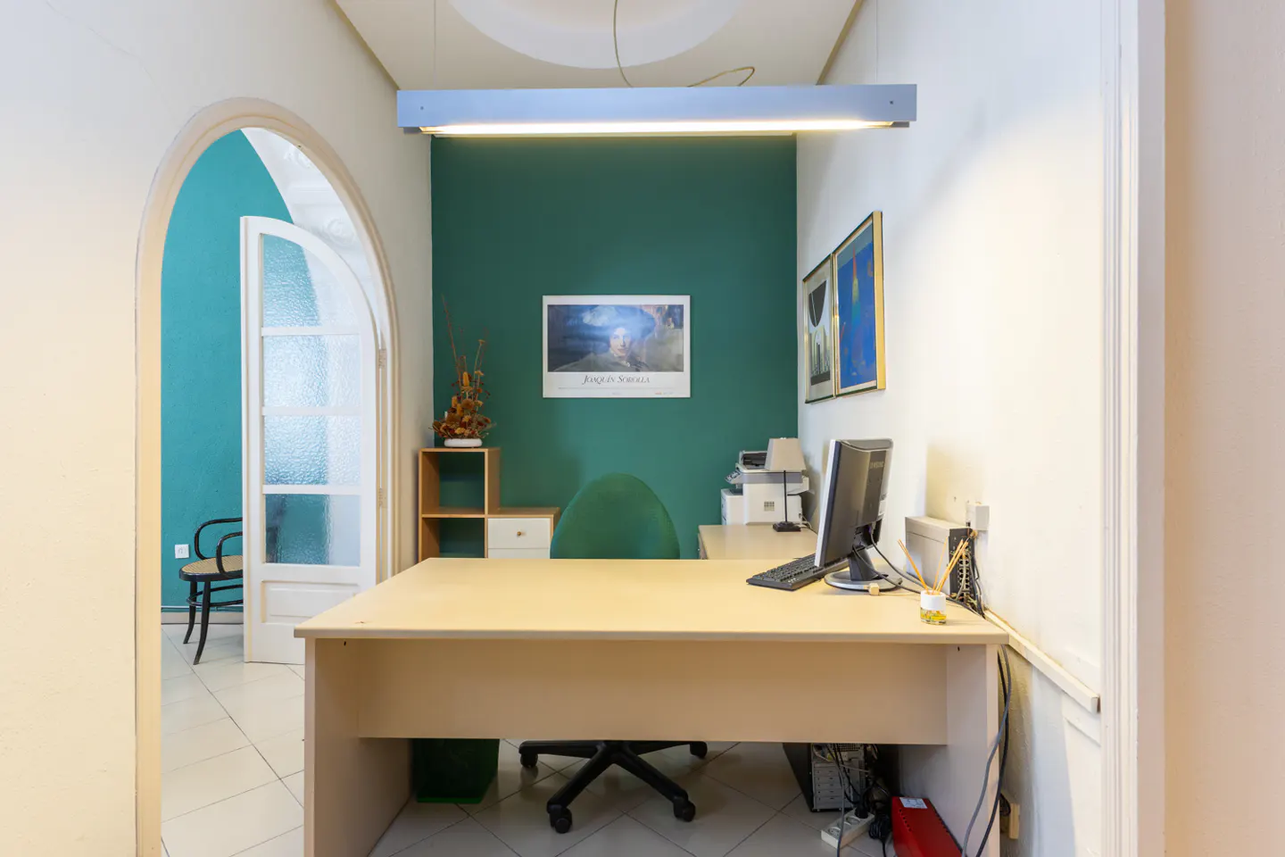 Office space with a light wood desk, computer, and green accent wall. An arched doorway leads to another room with teal walls.