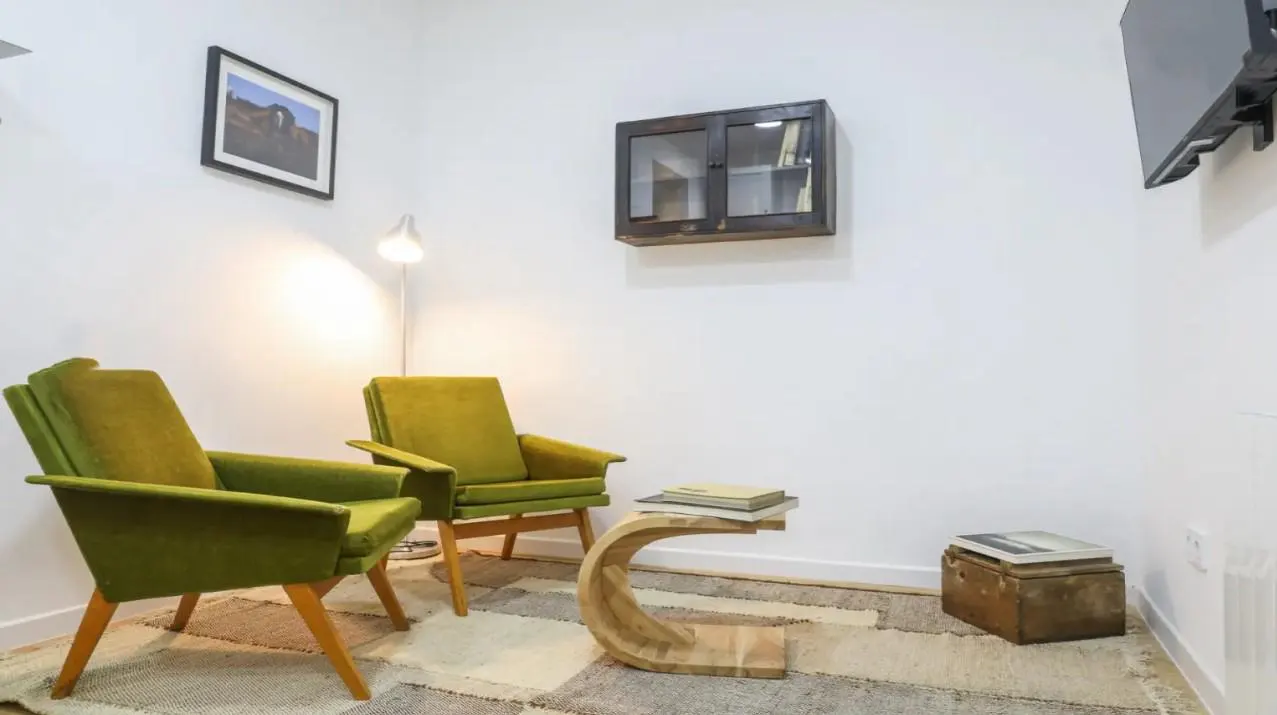 Living room with two green armchairs, a wooden table with books, and a patterned rug on a white background.