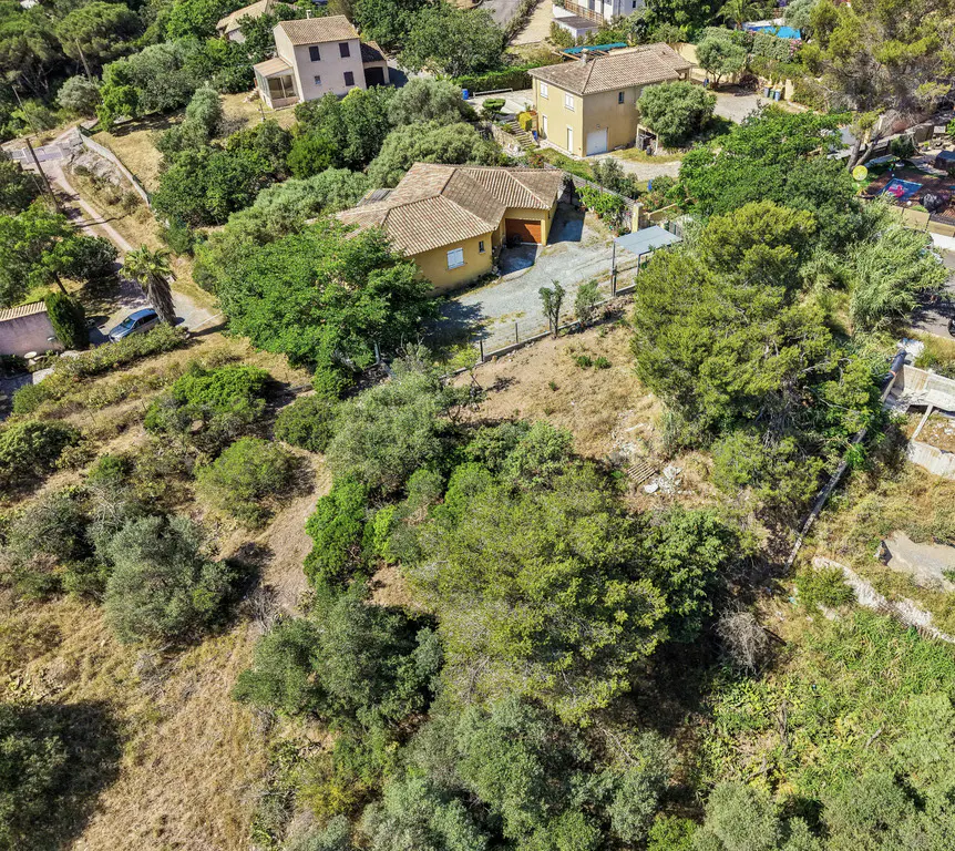 Aerial view of a tan villa with a tiled roof surrounded by lush green trees and dry, grassy terrain in a sunny landscape.