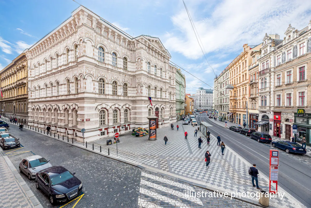 Street view of Prague, Czech Republic, with ornate buildings, cars, and pedestrians on a sunny day.