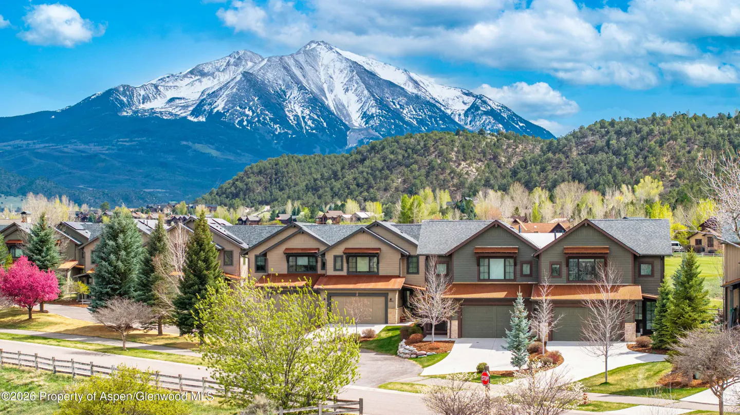 View of townhouses with garages, green lawns, and trees, set against a backdrop of mountains and a blue sky.