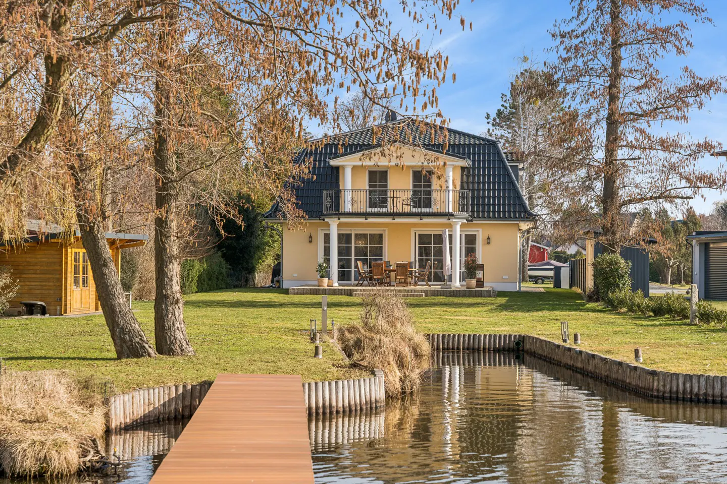 A yellow two-story house with a black roof sits on a grassy lawn next to a canal with a wooden dock.