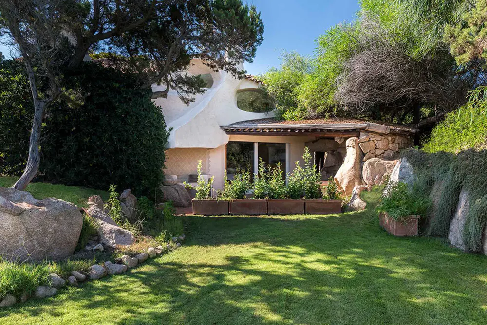 Exterior view of a white stucco house with stone accents, surrounded by lush greenery and a green lawn.